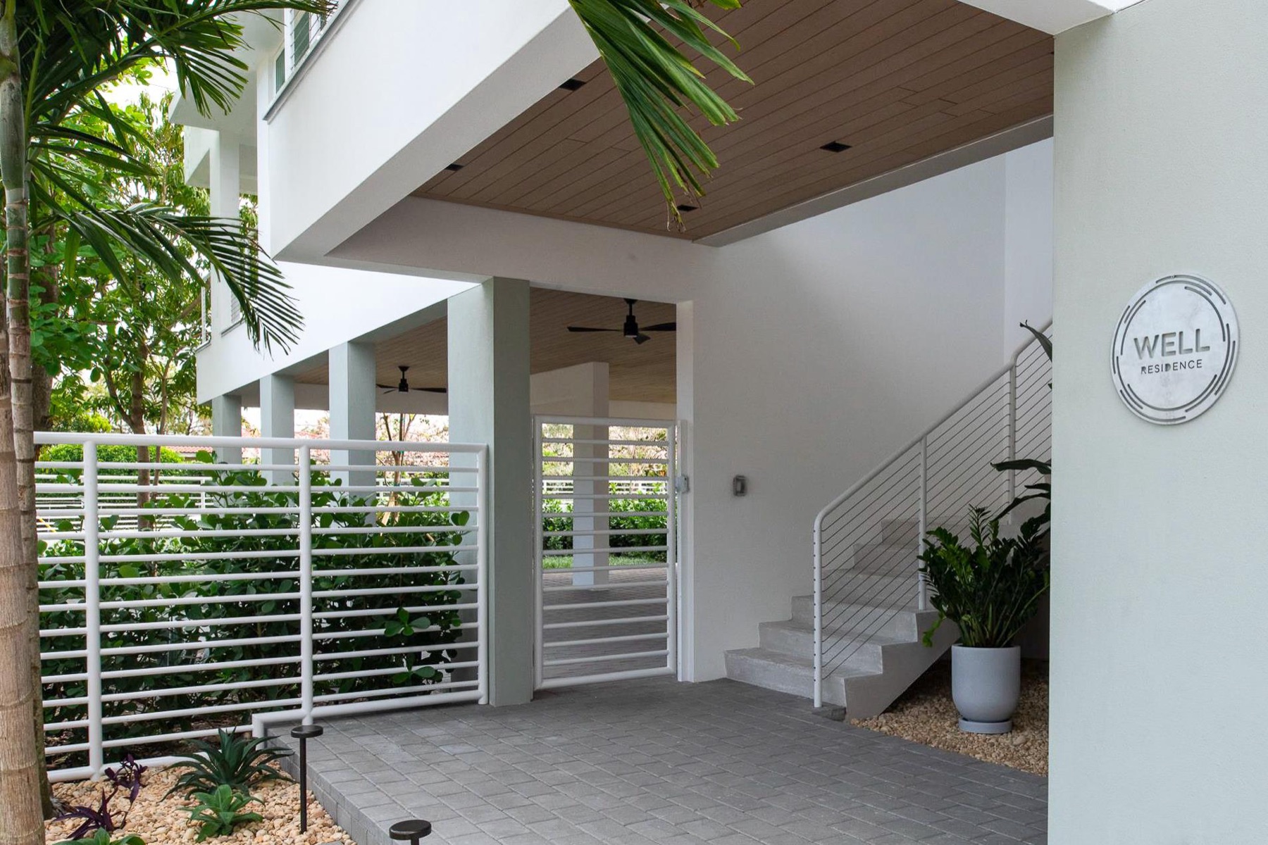 Entry courtyard beneath CM1's elevated living level, with an exterior stair, gate, and WELL Residence plaque on the wall