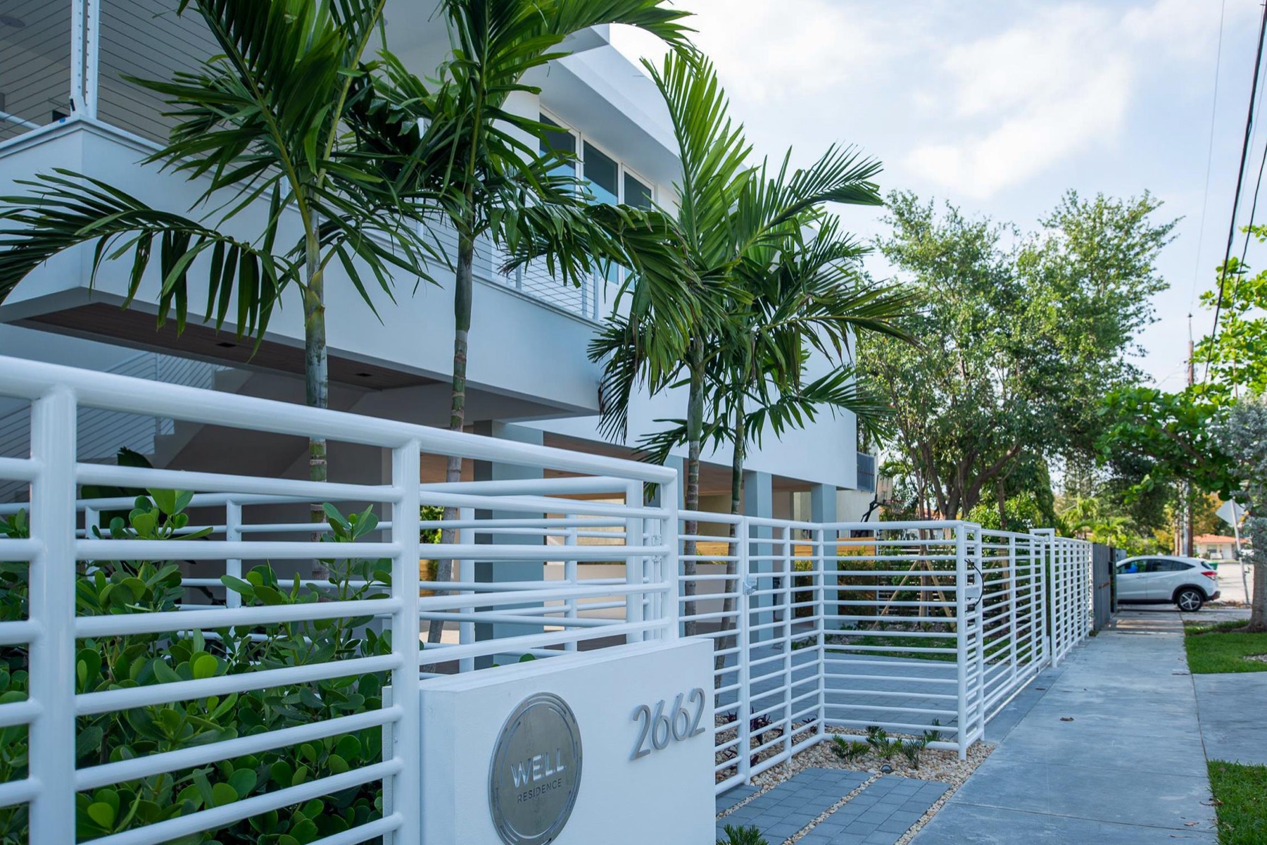Street view of CM1 with its white horizontal-bar fence, palm trees, and the elevated white volume above