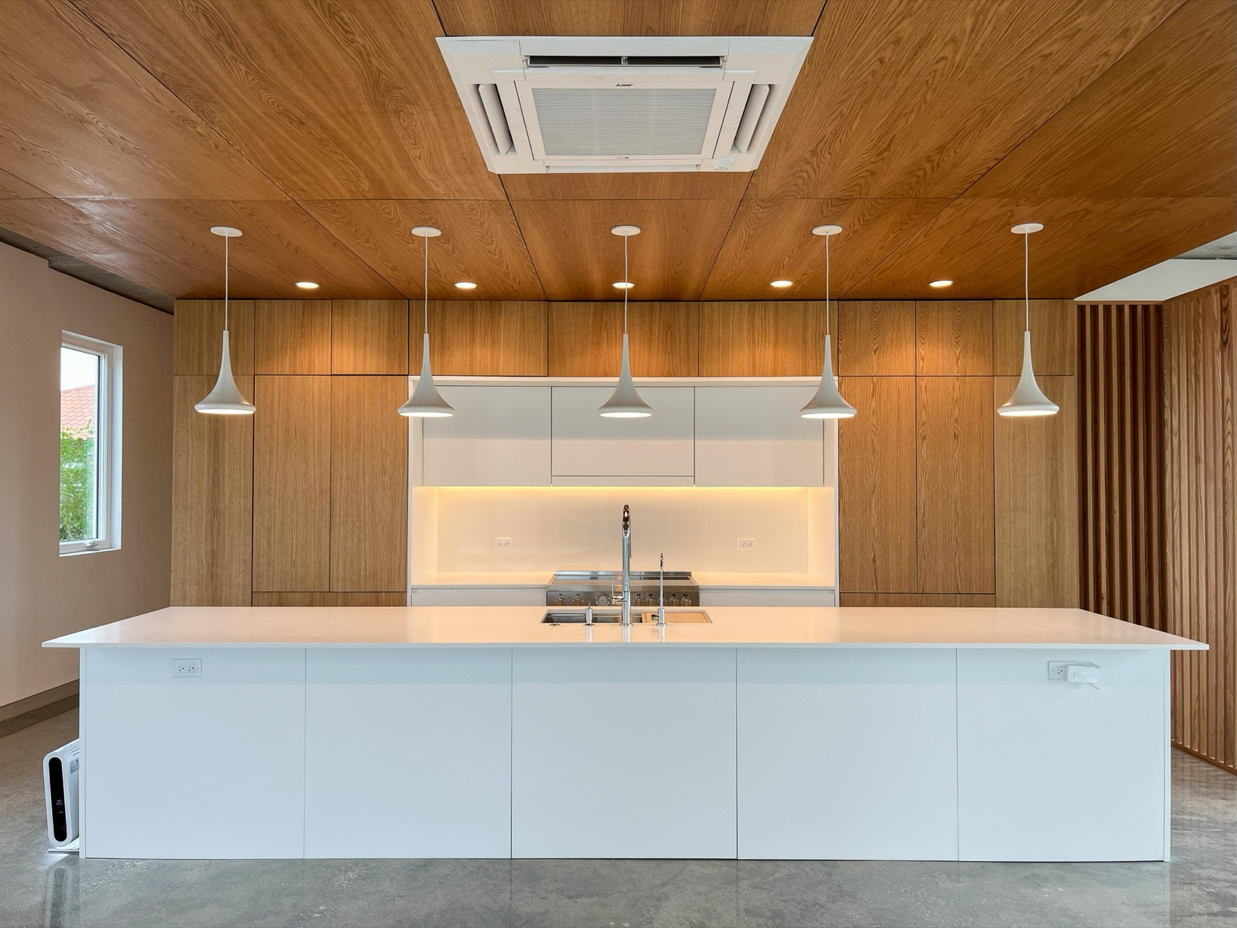 CM1's kitchen with a long white island, five white pendants, a ceiling cassette air-conditioning unit, and full-height oak paneling
