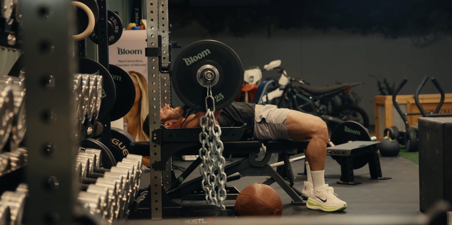 Greg LaVecchia bench pressing in a rack with chains hanging from the barbell, Bloom-stamped plates, dumbbells, kettlebells, and an electric dirt bike visible around the gym