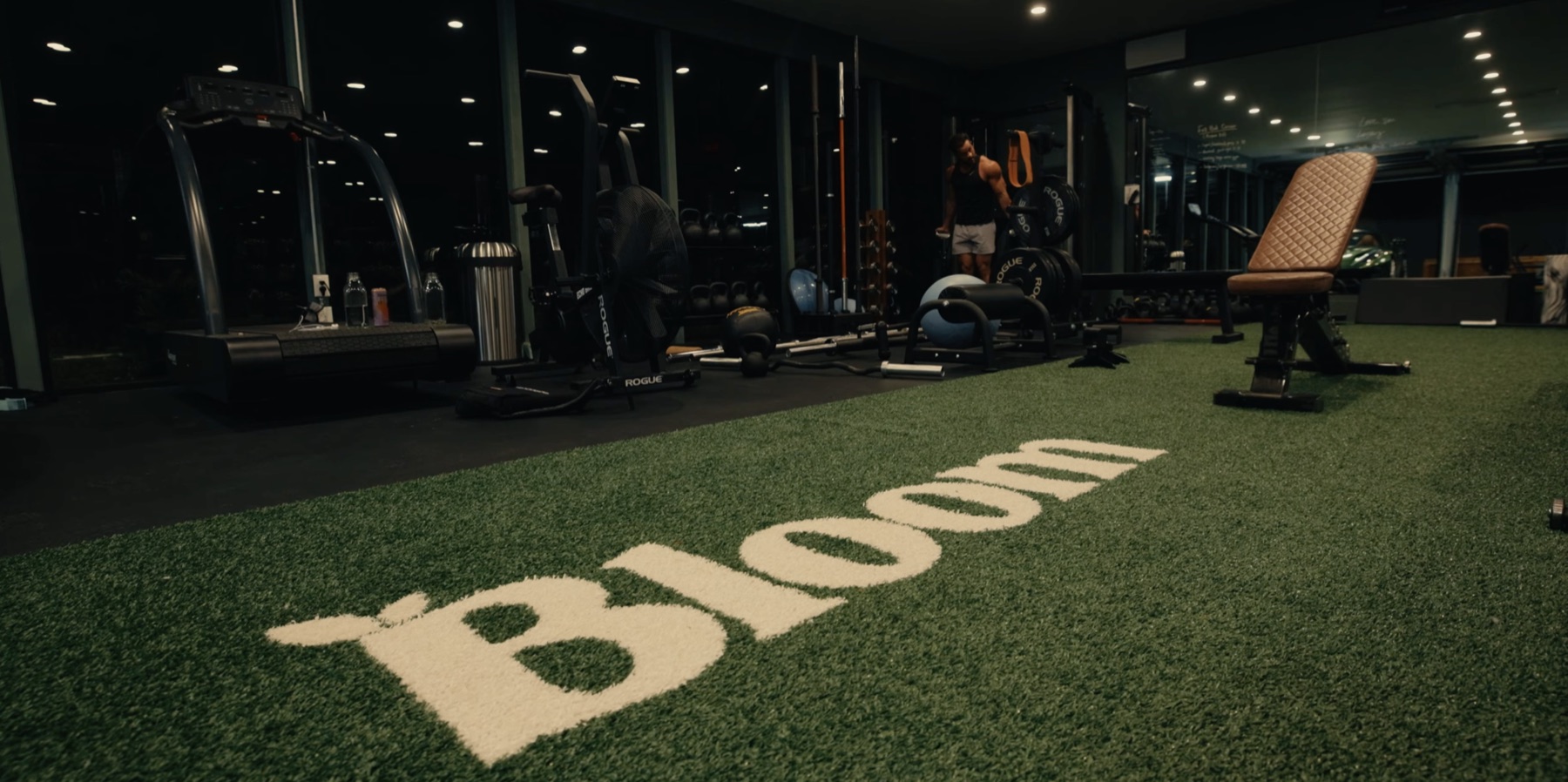 Low-angle view across a green turf lane with the Bloom logo inside Greg LaVecchia's gym, with a treadmill, Rogue cardio equipment, benches, racks, and mirrored walls in the background