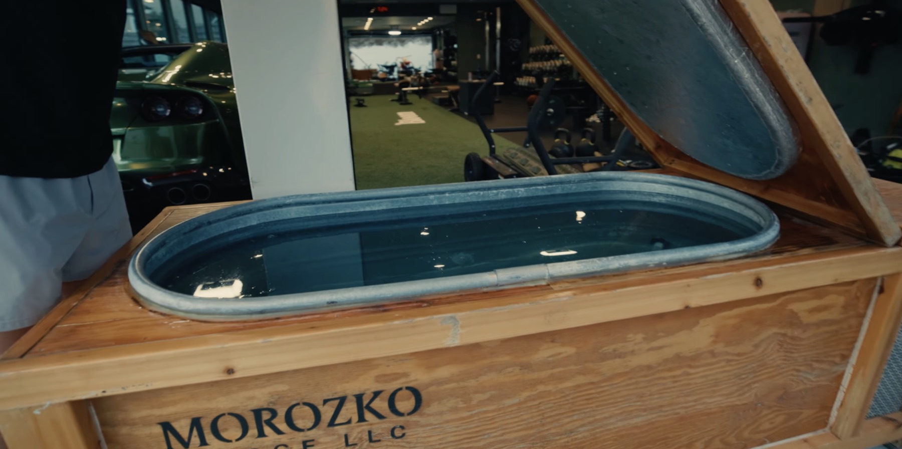 Close-up of a wooden Morozko Forge cold plunge with the lid open, positioned near Greg LaVecchia's gym with turf and equipment visible in the background