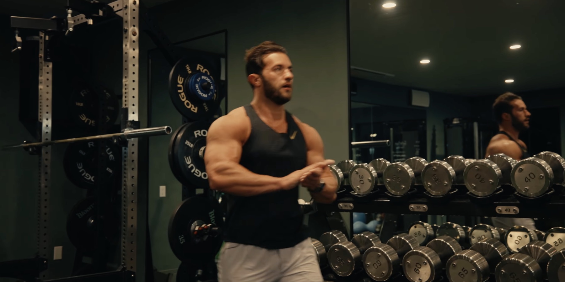 Greg LaVecchia standing beside a rack of polished custom dumbbells, with a squat rack, Rogue plates, mirrors, and green walls behind him