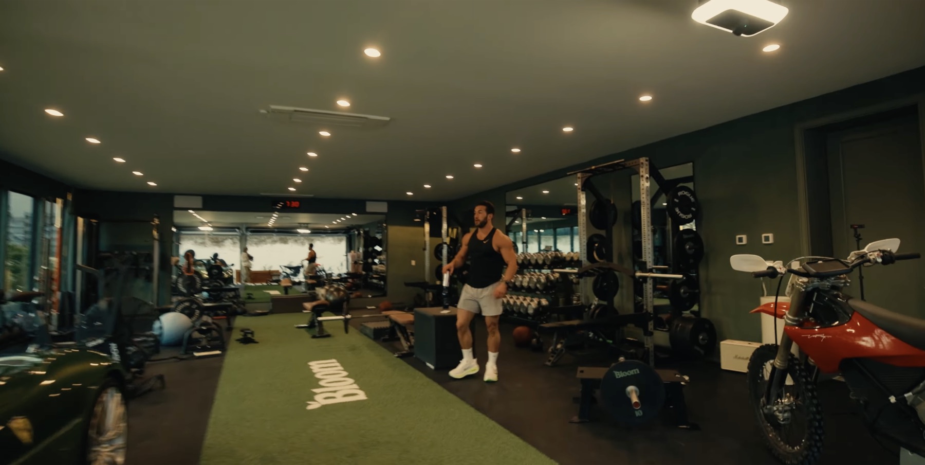 Wide view of the Bloom Gym showing a green Ferrari on the left, a red electric dirt bike on the right, turf down the center, racks, dumbbells, mirrors, and Greg standing in the middle