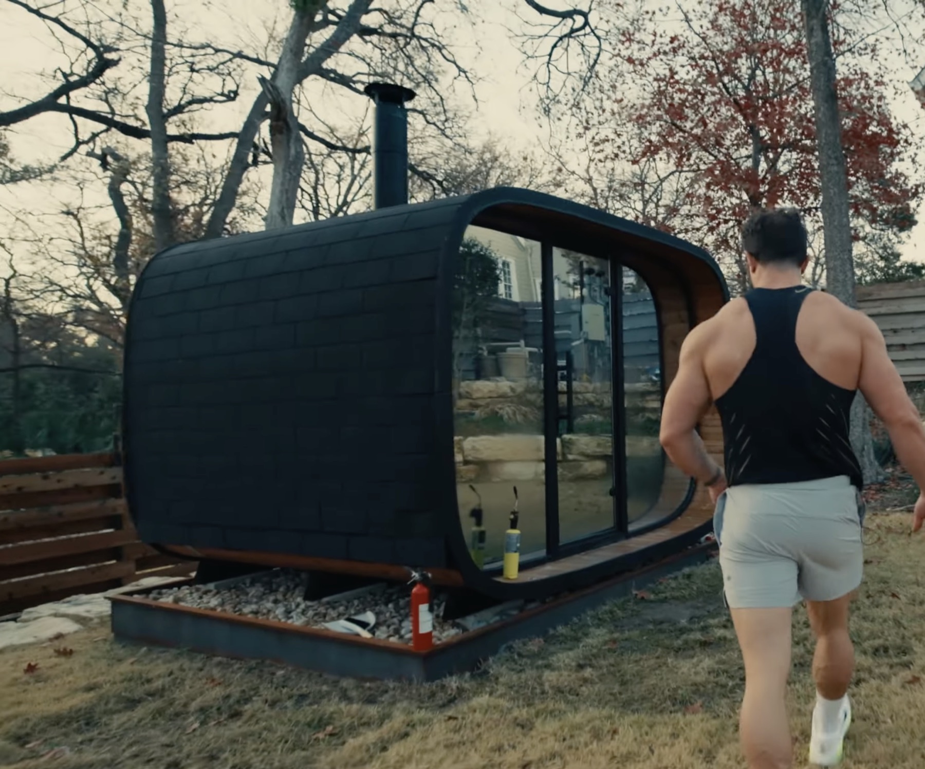 Greg LaVecchia walking toward a large black wood-burning outdoor sauna with glass front, rounded corners, trees, fence, and Austin backyard landscaping around it