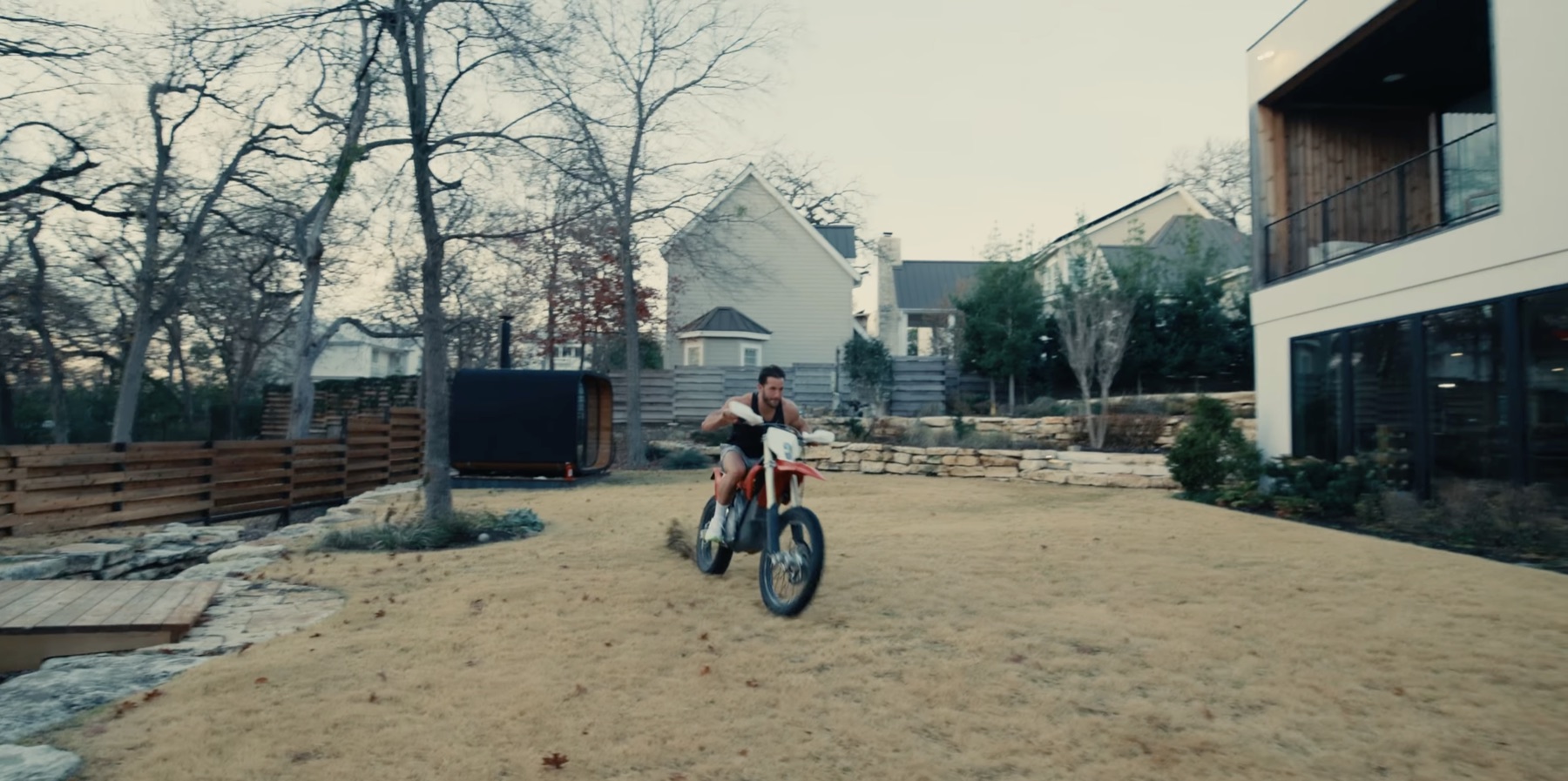 Greg LaVecchia riding a red dirt bike across a large Austin backyard with a black sauna, trees, fence, stone landscaping, and the gym building visible nearby