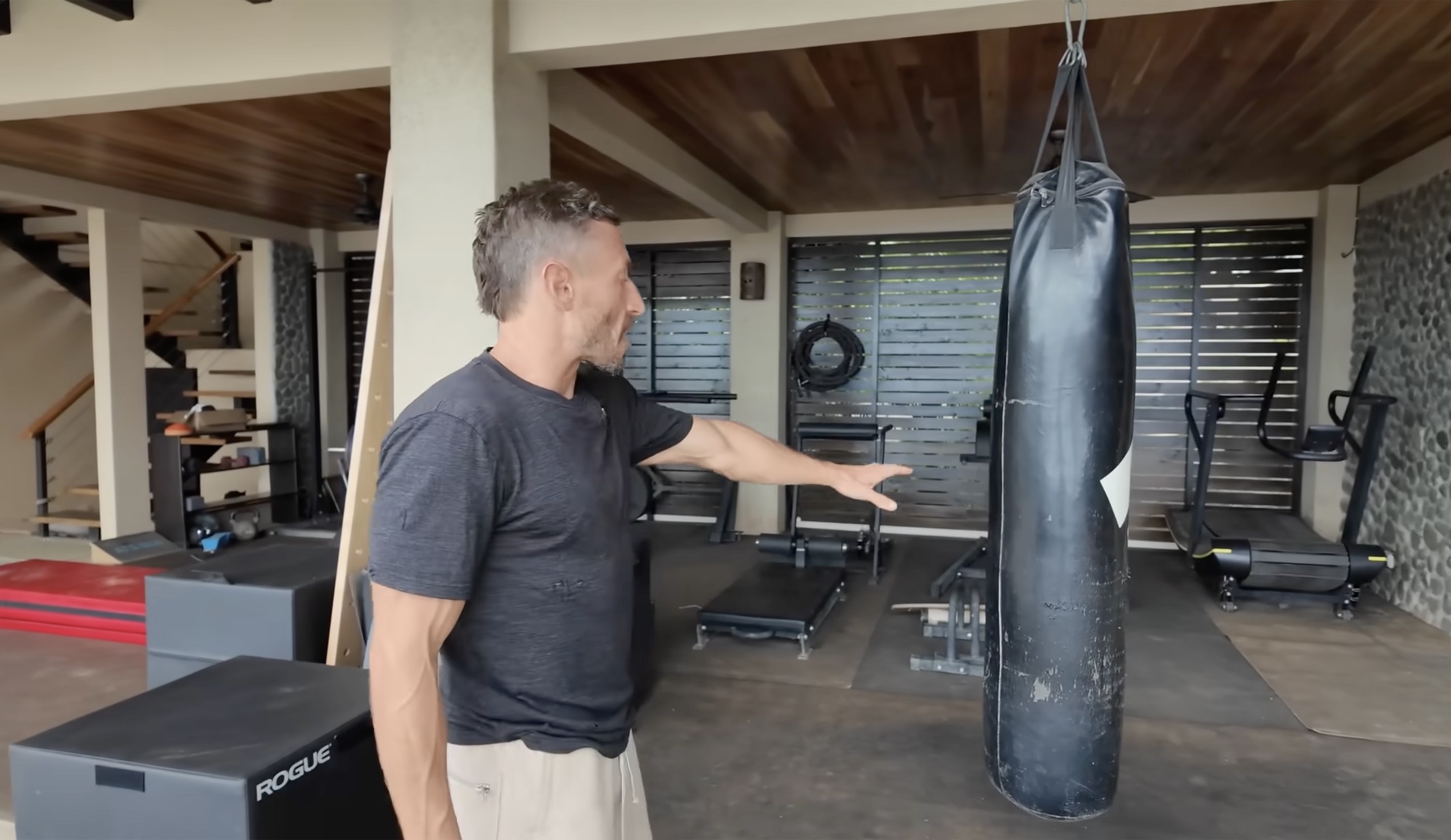 Covered outdoor gym — Paul pointing at a well-worn heavy bag, Rogue box jump, Nordic bench, and two treadmills under a wood-slat ceiling