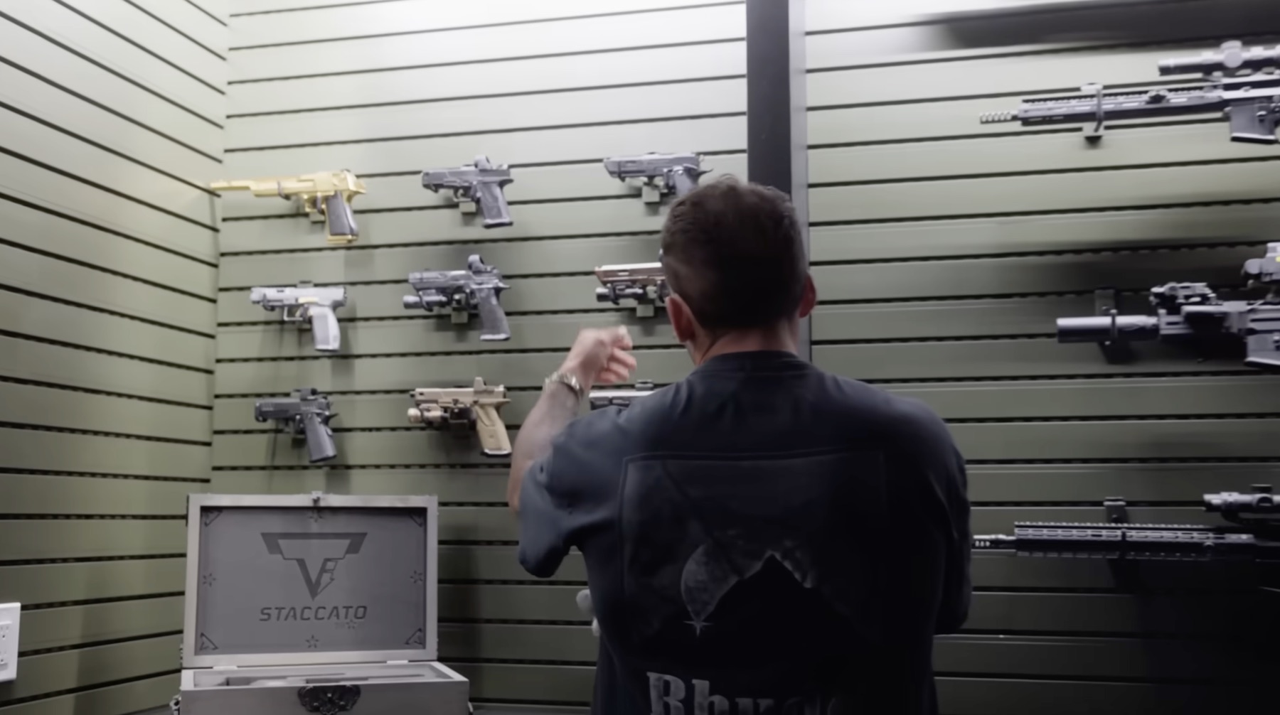 Dom Iacovone standing in front of a green slat-wall armory with pistols and rifles mounted on display rails
