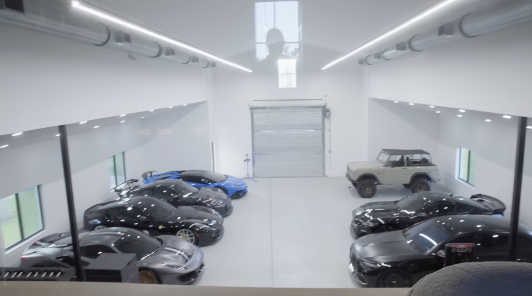 High-angle view inside Dom Iacovone's bright white garage with multiple sports cars parked on both sides, a roll-up door at the far end, and linear lights running across the ceiling