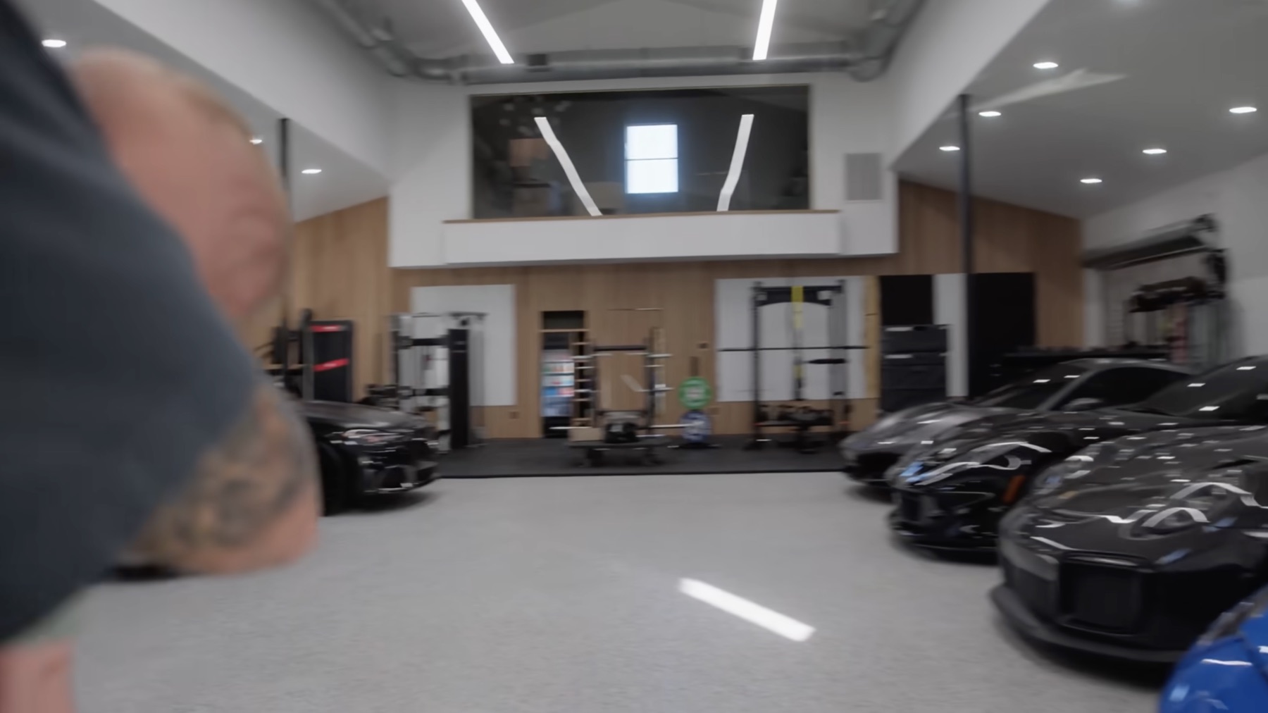 Ground-level view of Dom Iacovone's garage with sports cars parked along both sides, gym equipment and wood slat walls in the background, and an upstairs loft visible above