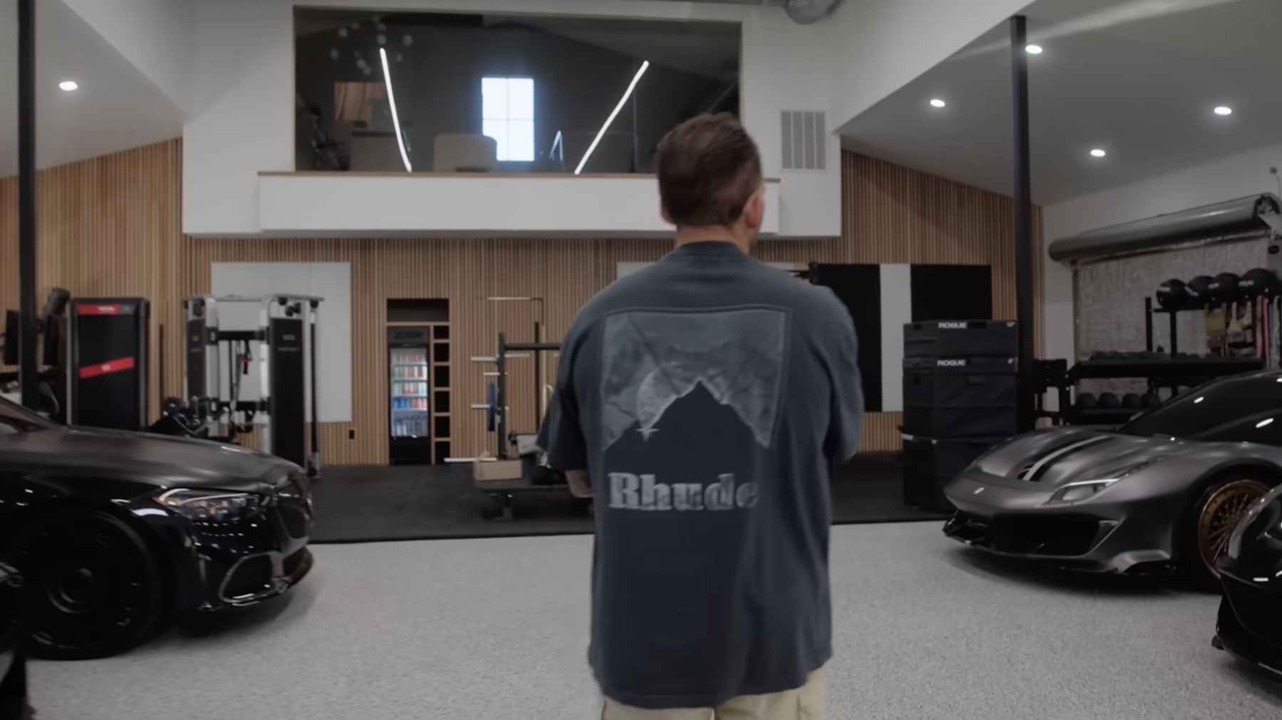 Wide view of the gym zone inside Dom Iacovone's garage, with cars on both sides, wood slat walls, cardio and strength equipment, and a glass office loft overhead