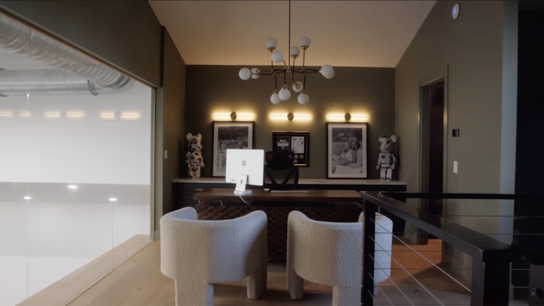 Upstairs loft office in Dom Iacovone's garage with olive-green walls, warm wall sconces, framed awards and articles, a wood desk, cream chairs, and a view down into the garage on the left
