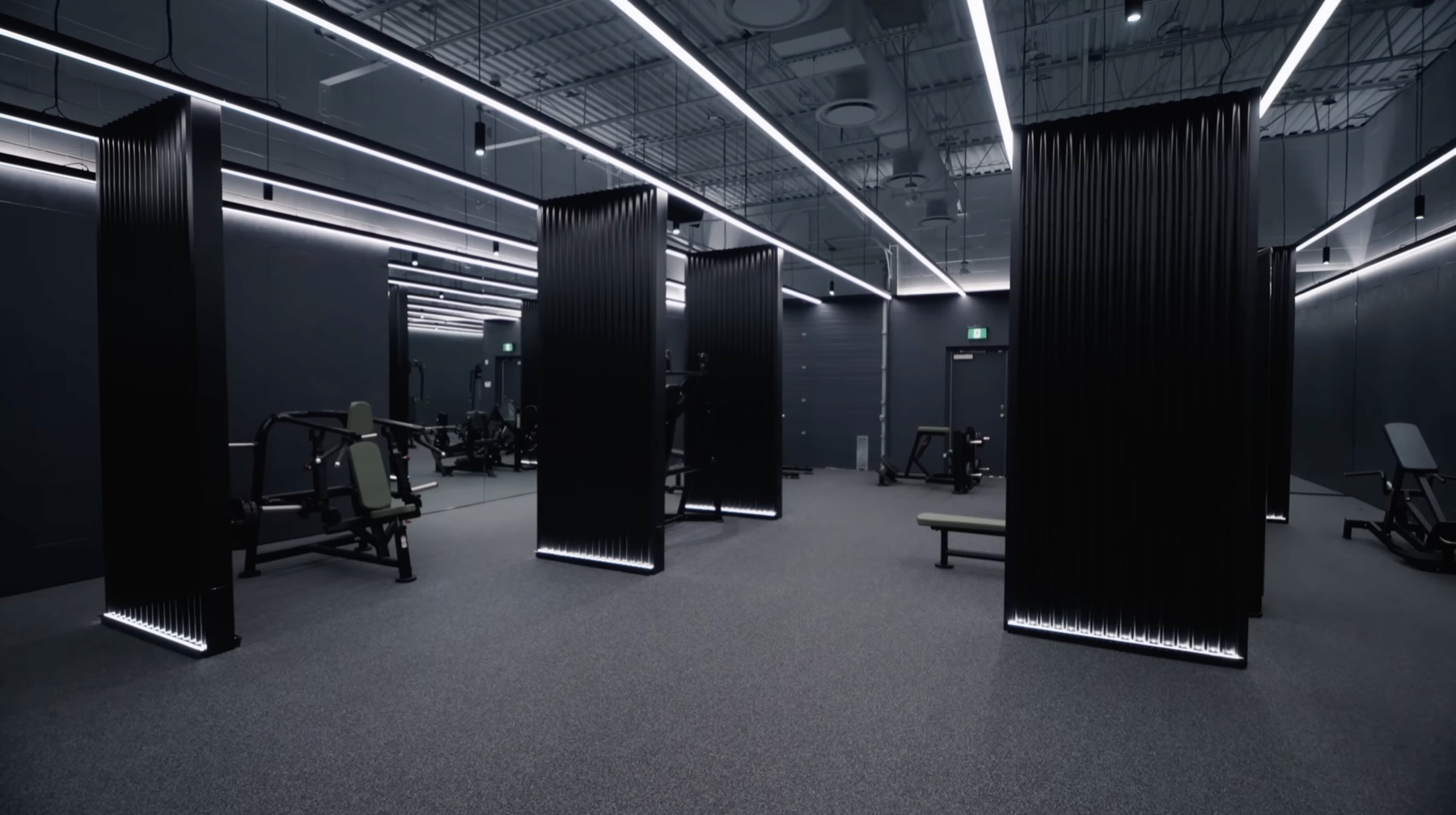 The dark gym — tall black fluted columns with vertical white LED strips on their edges, spaced throughout a dark-walled room with black rubber flooring and machines partially visible between them, everything lit dramatically from above