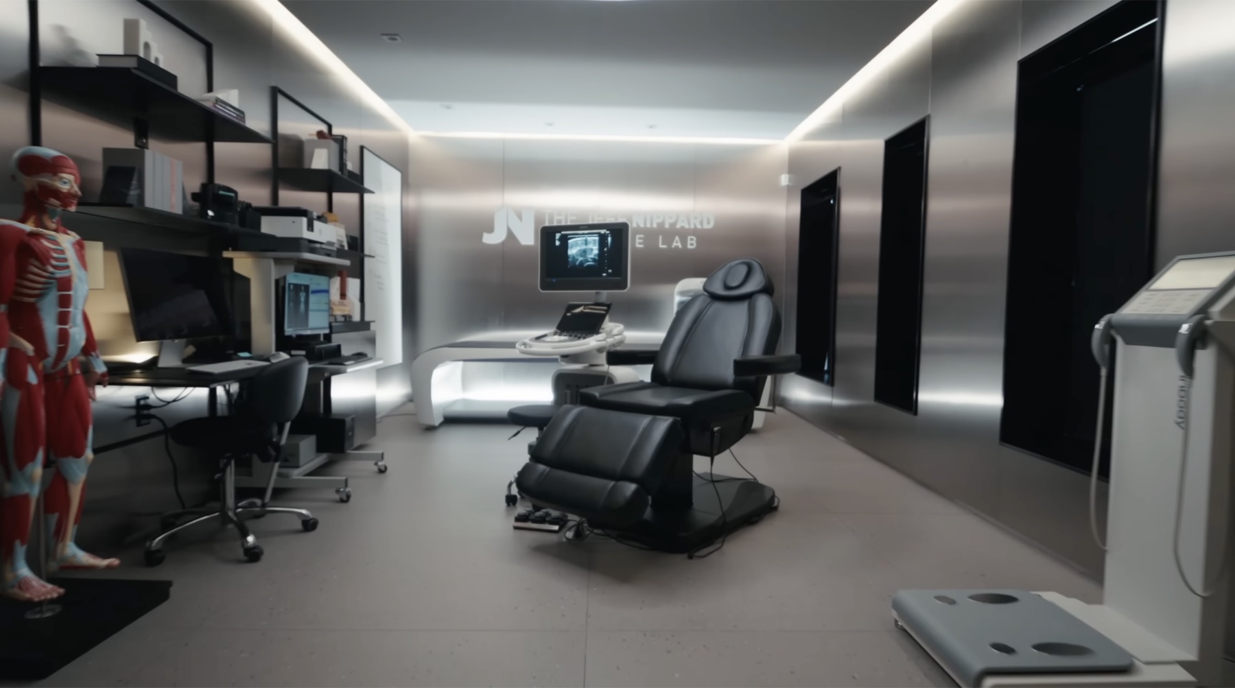 The Muscle Lab research room — anatomical muscle model on the left, desk workstations with monitors, a black adjustable examination chair in the center, an ultrasound machine on a rolling stand, brushed-steel walls with 'THE JEFF NIPPARD MUSCLE LAB' signage, BIA scale in the foreground right, EMG cabinet on the far right