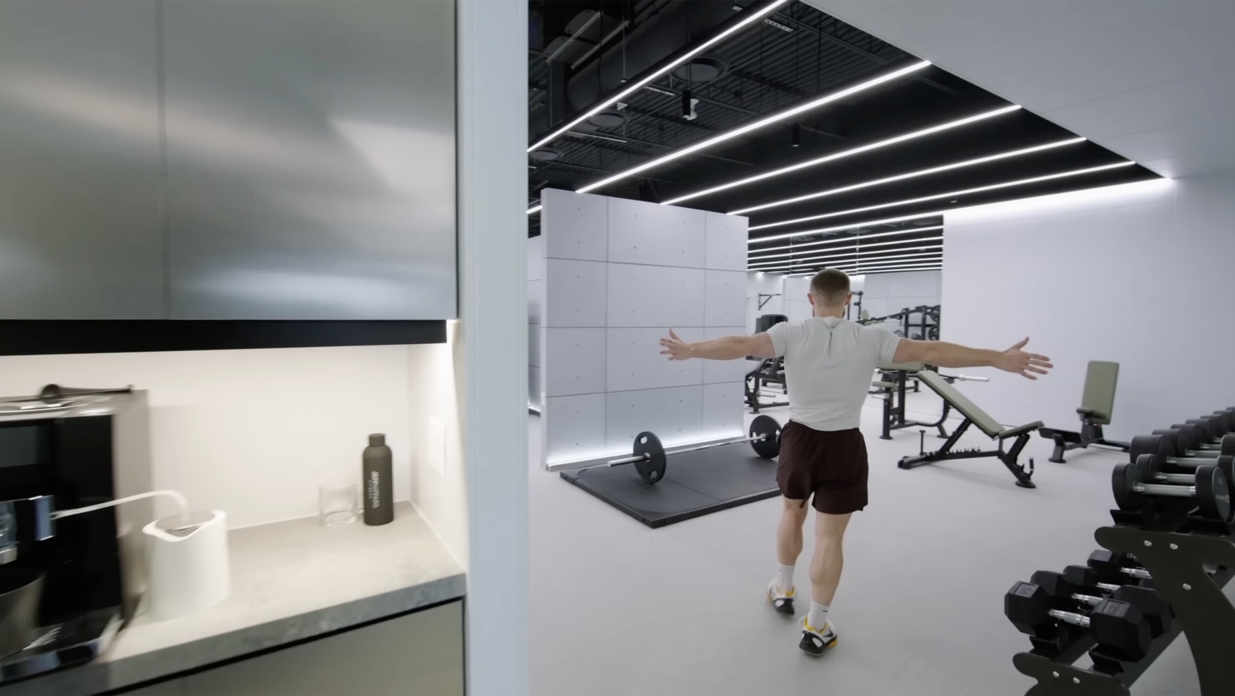 Jeff Nippard photographed from behind with arms spread, walking from a kitchen area into a bright white gym with linear ceiling lights, a deadlift platform with a loaded barbell, an adjustable bench, a squat rack, and dumbbell racks along the right wall