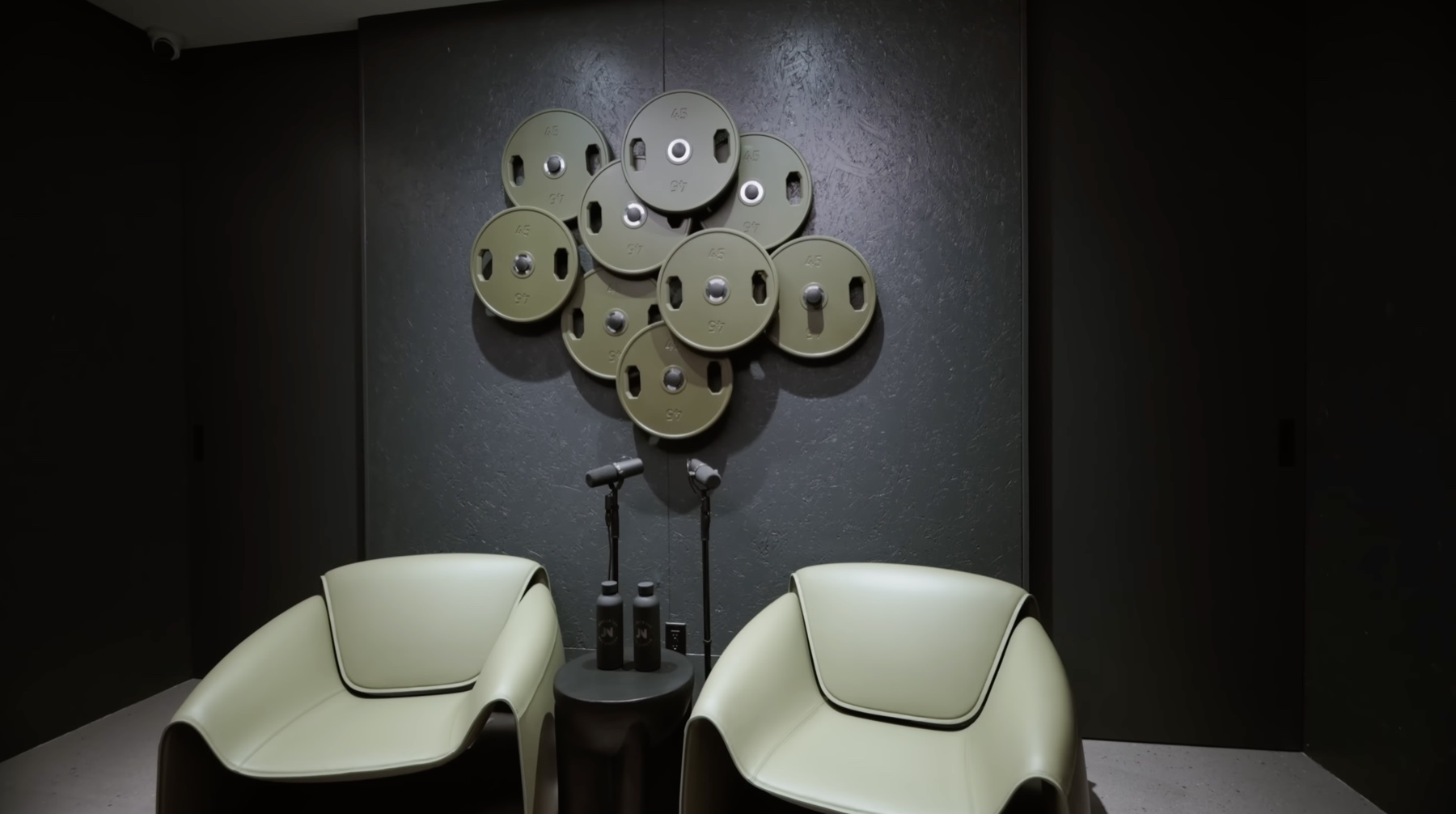 The Muscle Lab lobby — dark walls with a cluster of green 45lb plates arranged in a flower pattern as wall art, two cream-colored armchairs facing each other across a small round side table with dumbbells on top