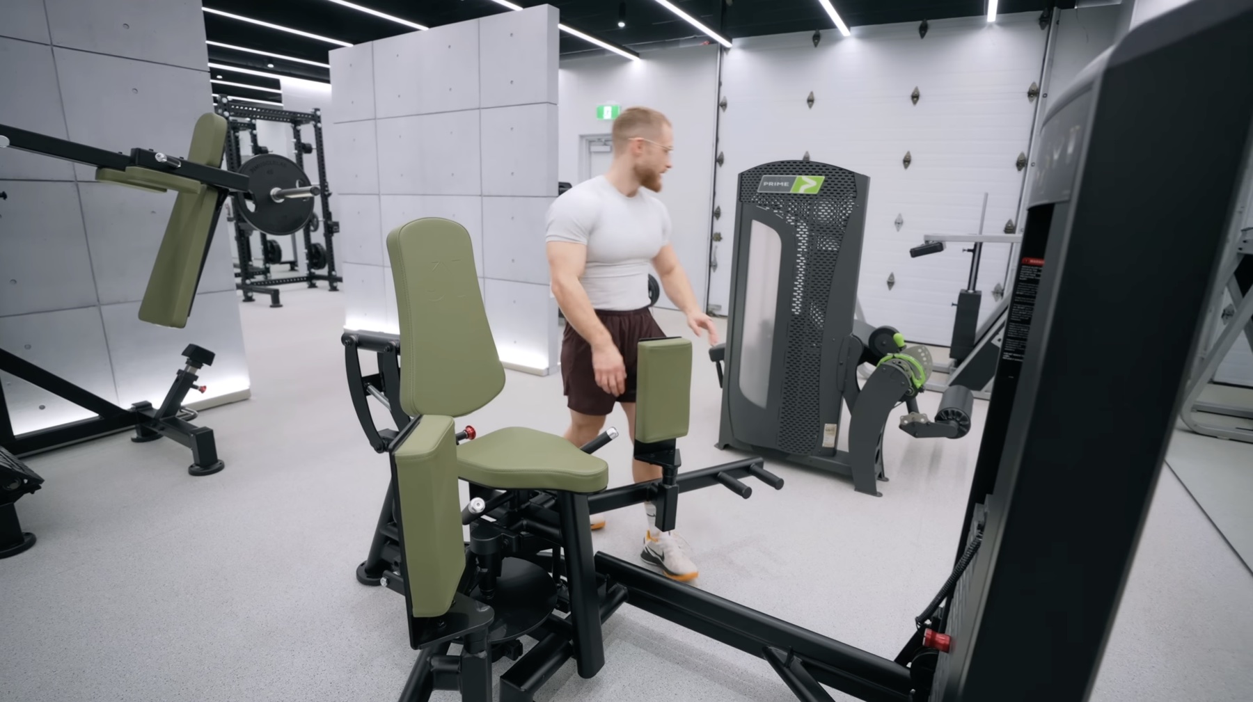 Jeff standing next to a Prime hip abduction and adduction machine with olive-green padded seats, a squat rack with plates loaded in the background and other machines in the bright white room