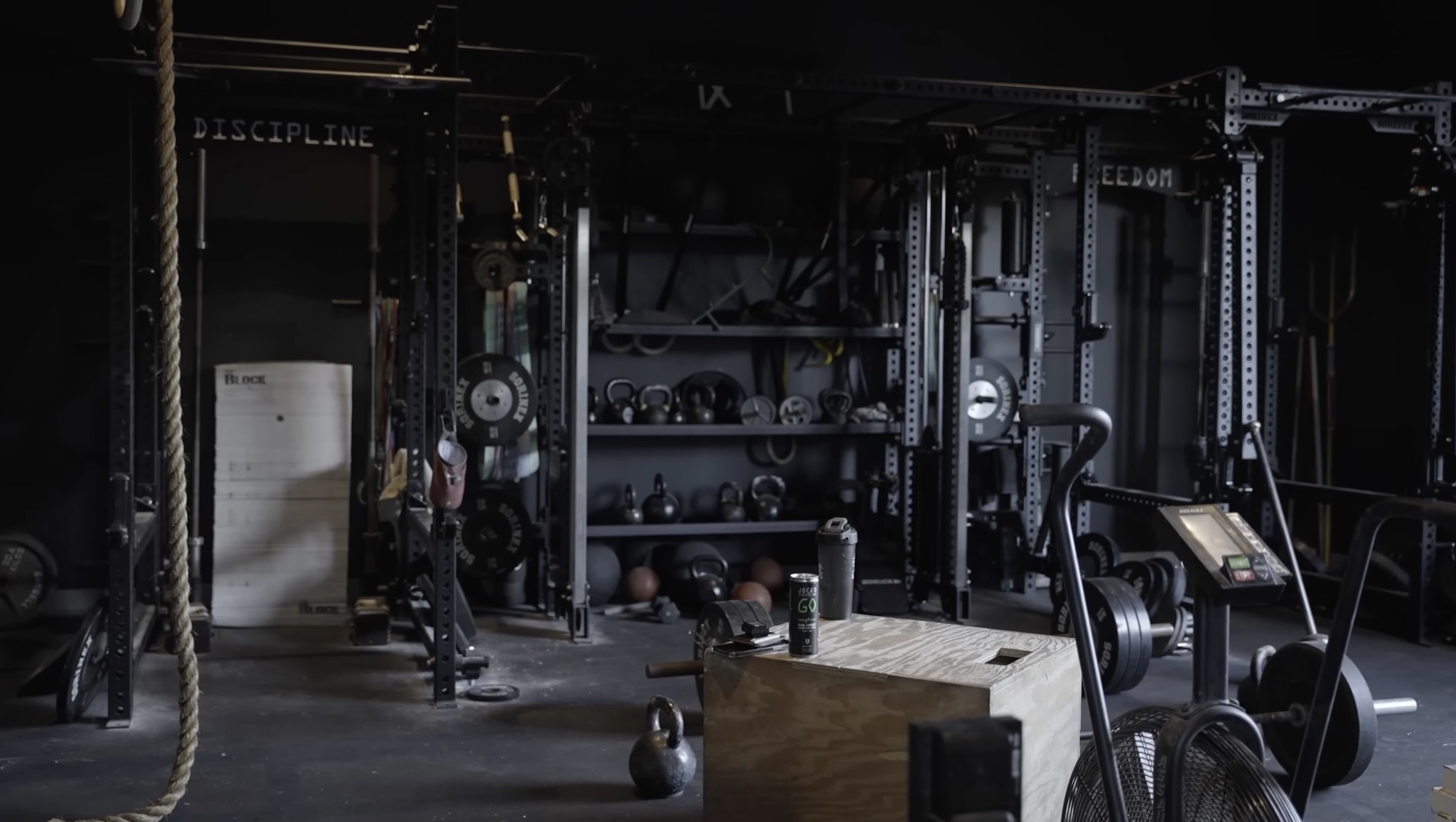 Wide interior shot of Jocko Willink's home gym — a Sorinex rig with 'DISCIPLINE' and 'FREEDOM' stencilled across the uprights, plate storage, kettlebell shelves, a climbing rope, plyo box, and an Assault Bike under low warehouse lighting