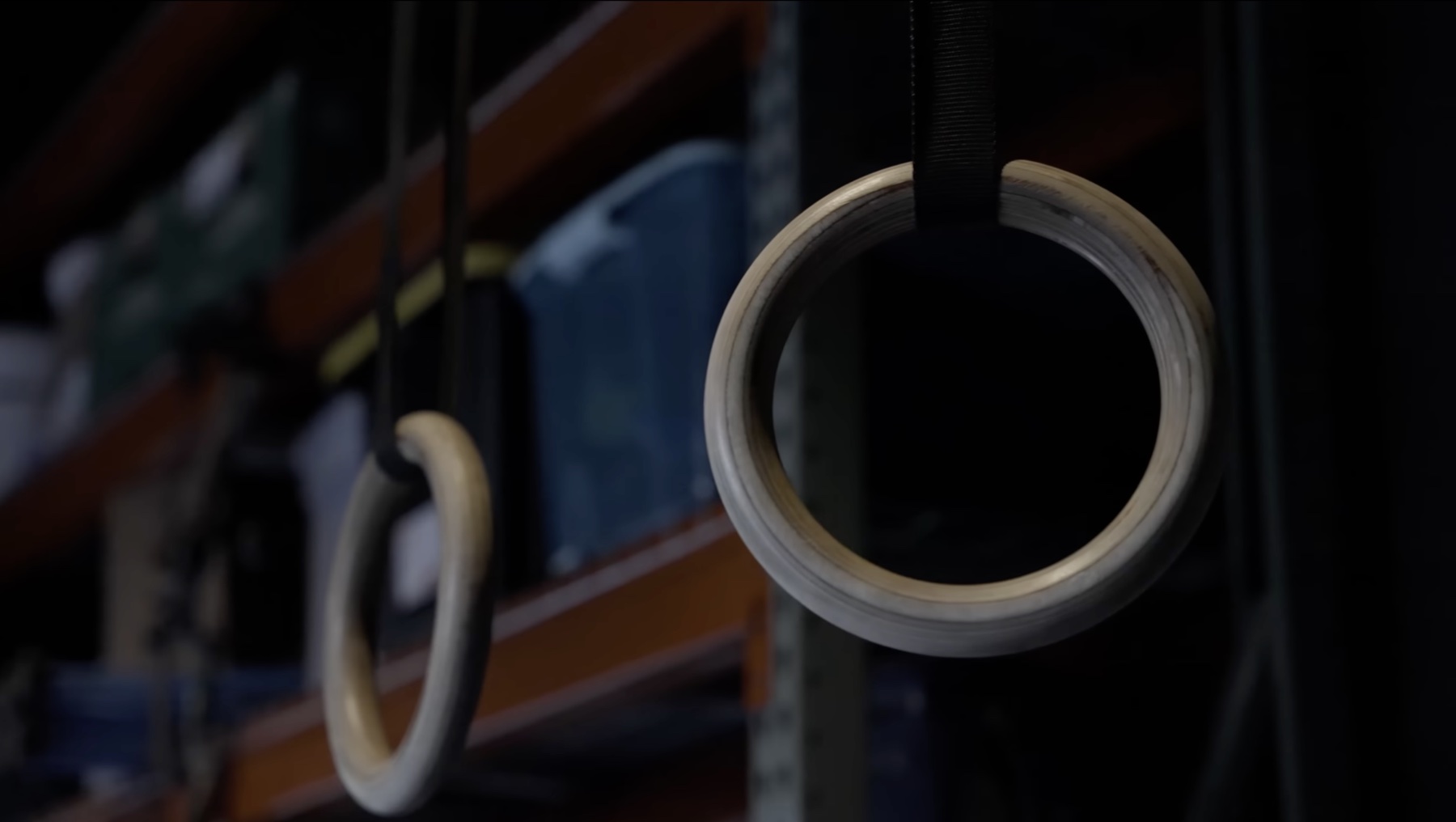 Close-up of two wooden gymnastic rings hanging on black straps in front of a dark plate rack, one ring in sharp focus, the other softly blurred in the foreground