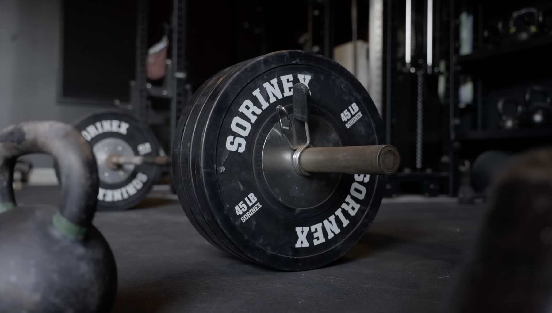 A loaded Sorinex barbell with three 45-pound black bumper plates stamped with the Sorinex logo, resting on a dark gym floor next to a kettlebell, with a second barbell and rack out of focus behind it