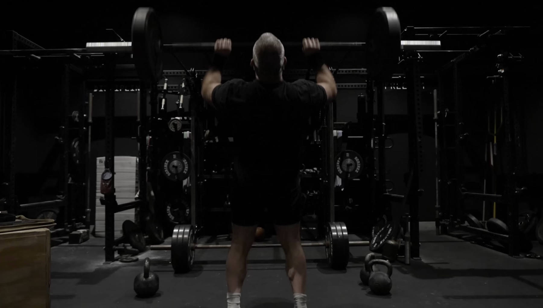 Jocko photographed from behind inside the rig, gripping a loaded barbell at shoulder height on the pins, kettlebells and plate storage visible on either side