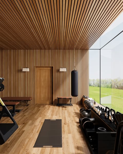 Oak-slatted home gym with warm wood flooring, a yoga mat, PENT dumbbells, and a hanging heavy bag beside a floor-to-ceiling window onto the countryside
