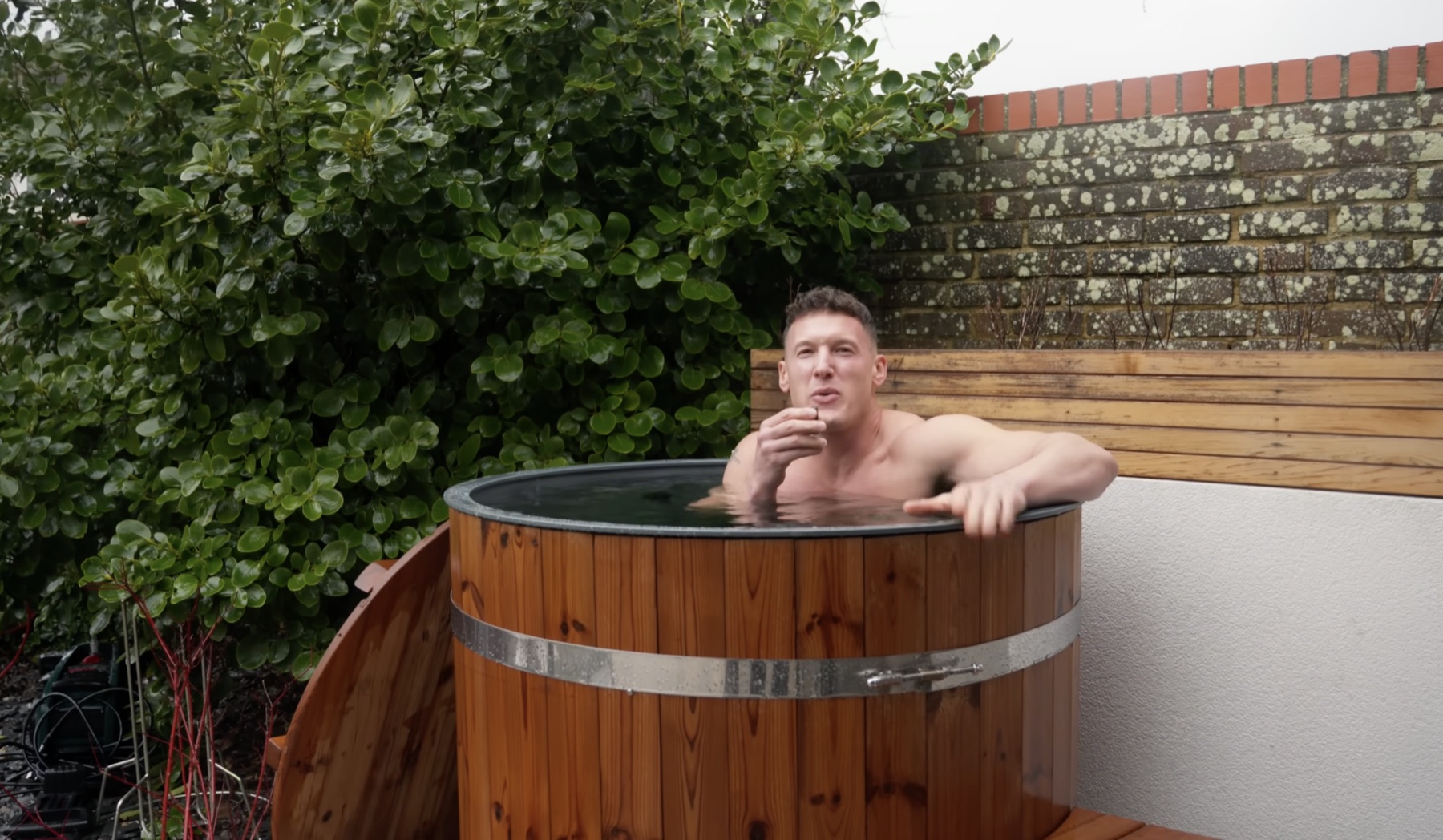 Matt in a cedar-slatted cold plunge tub against a brick wall with green foliage behind him, outdoor, horizontal wood slat privacy screen on the right