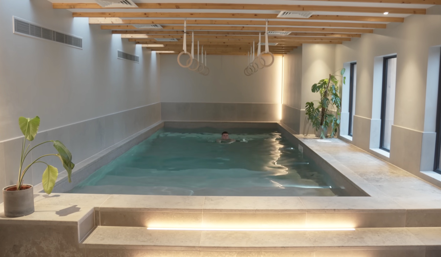 Glass-enclosed indoor lap pool in a long room with exposed oak beam ceiling, gymnastics rings hanging from the beams, stone surround, a plant in a concrete pot in the foreground, Matt mid-swim