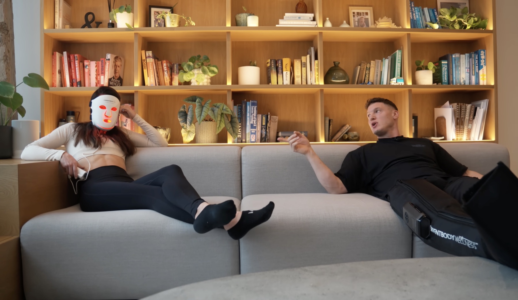Matt and Sarah on a grey sofa in a warm bookshelf-lined living room — Sarah wearing a CurrentBody red-light face mask on the left, Matt wearing CurrentBody compression boots on the right
