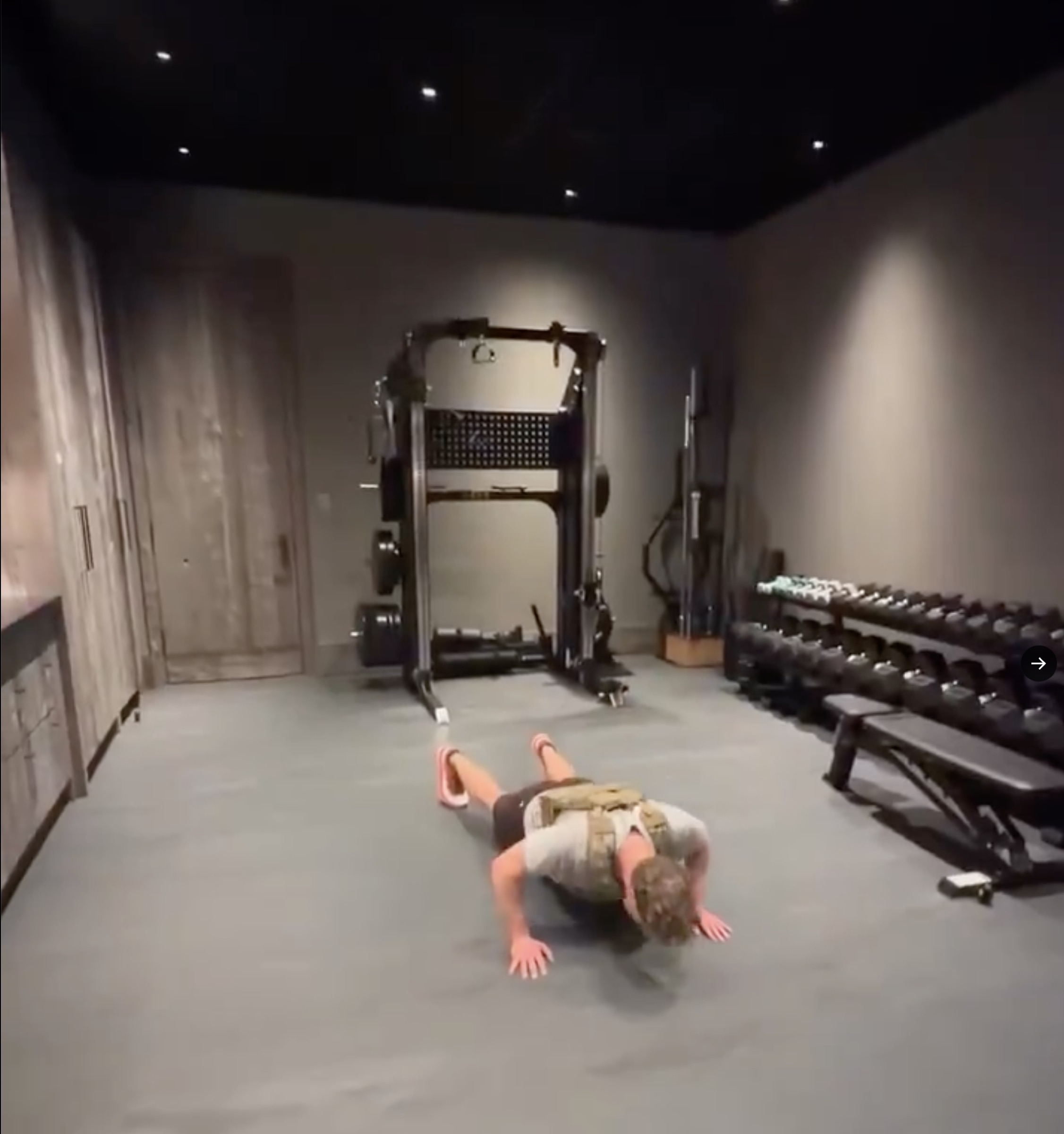 Wide interior of Mark Zuckerberg's home gym — a black integrated rack with plate storage and pegboard against a wood-paneled wall, a long row of hex-head dumbbells and an adjustable bench down the right side, polished concrete floor under a dark ceiling
