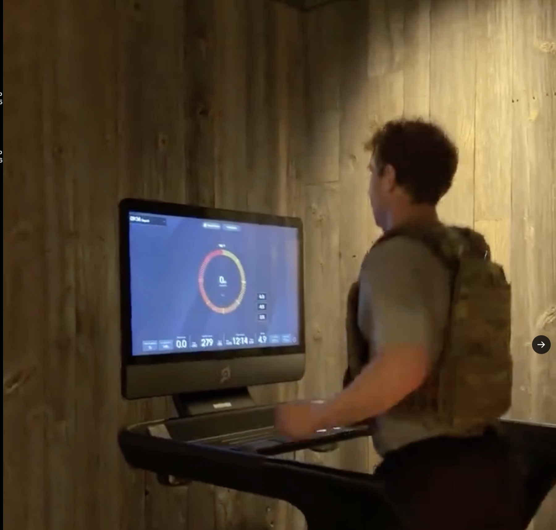 A trainee in a tactical weight vest walks on a Peloton Tread facing the touchscreen, against a warmly lit wood-paneled wall