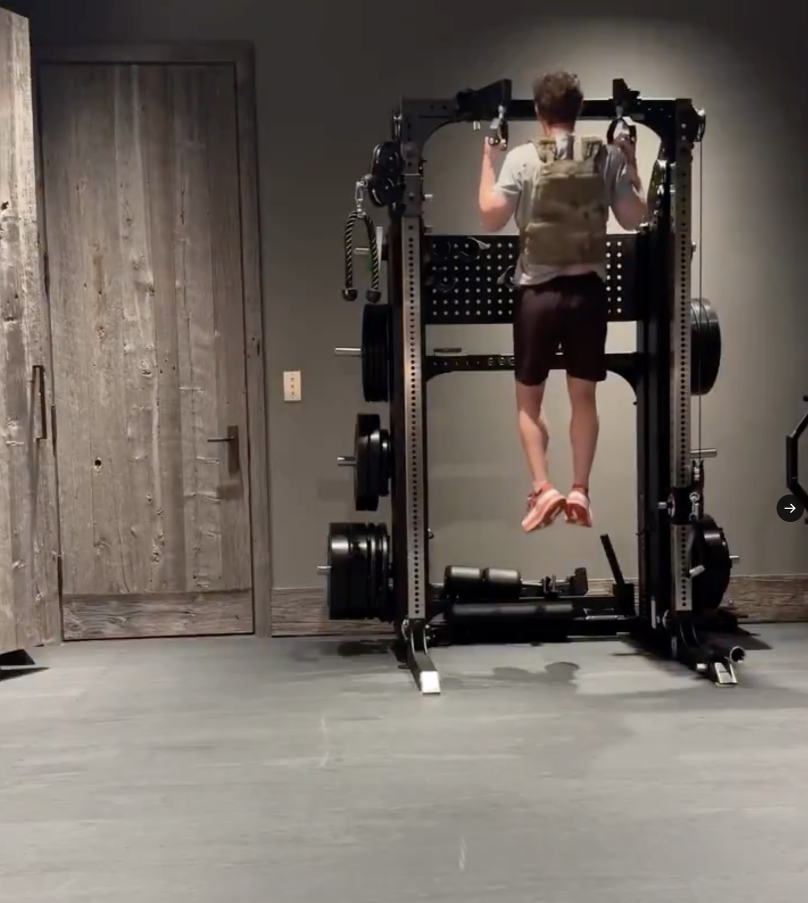 A trainee in a tactical weight vest performs weighted pull-ups on the black Eleiko Prestera rack, plate storage stacked down the uprights, a rustic wood door to the left