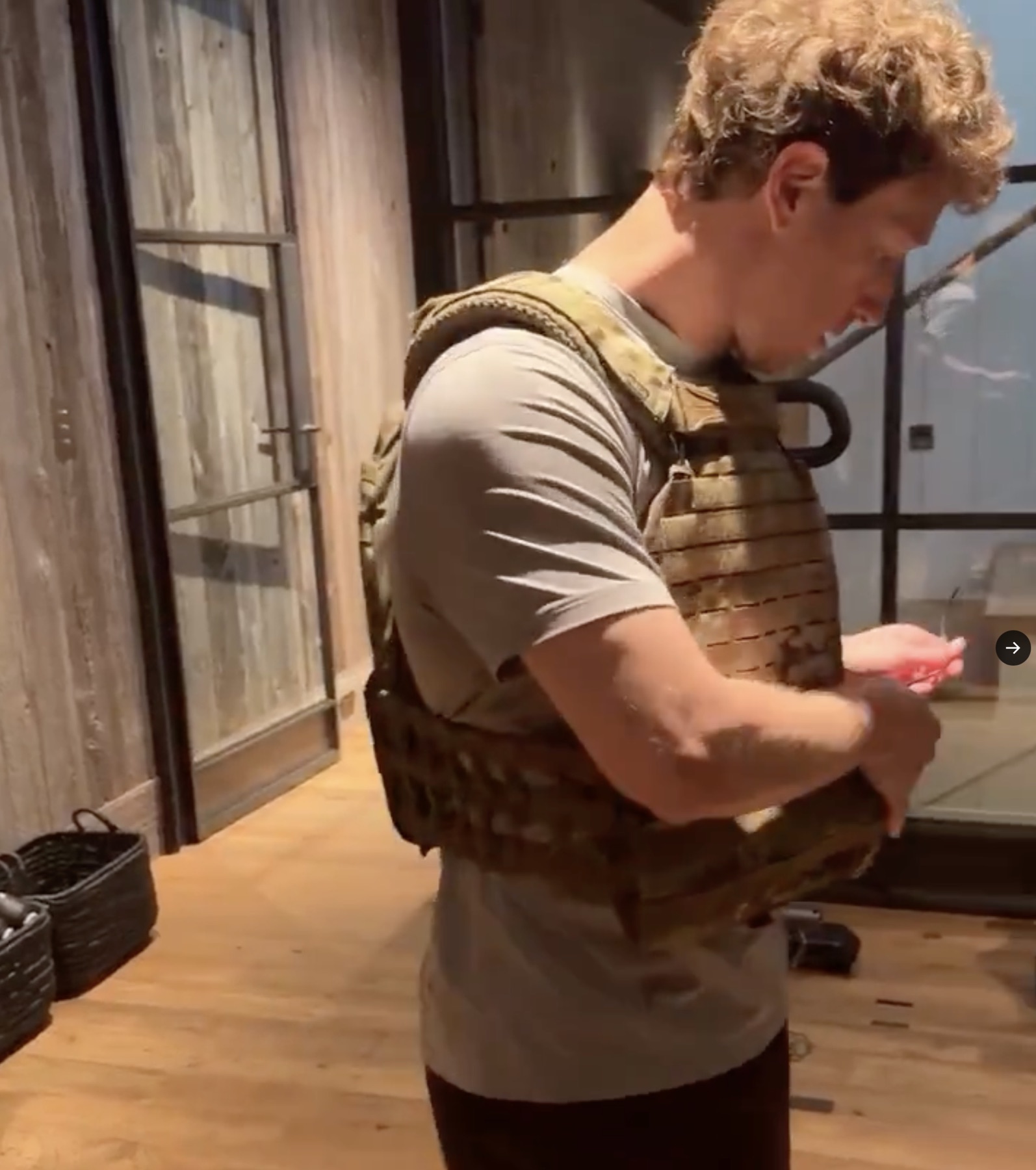 Close-up of Mark Zuckerberg adjusting a tan 5.11 Tactical plate carrier over a grey t-shirt, industrial-framed glass doors and wood paneling behind him